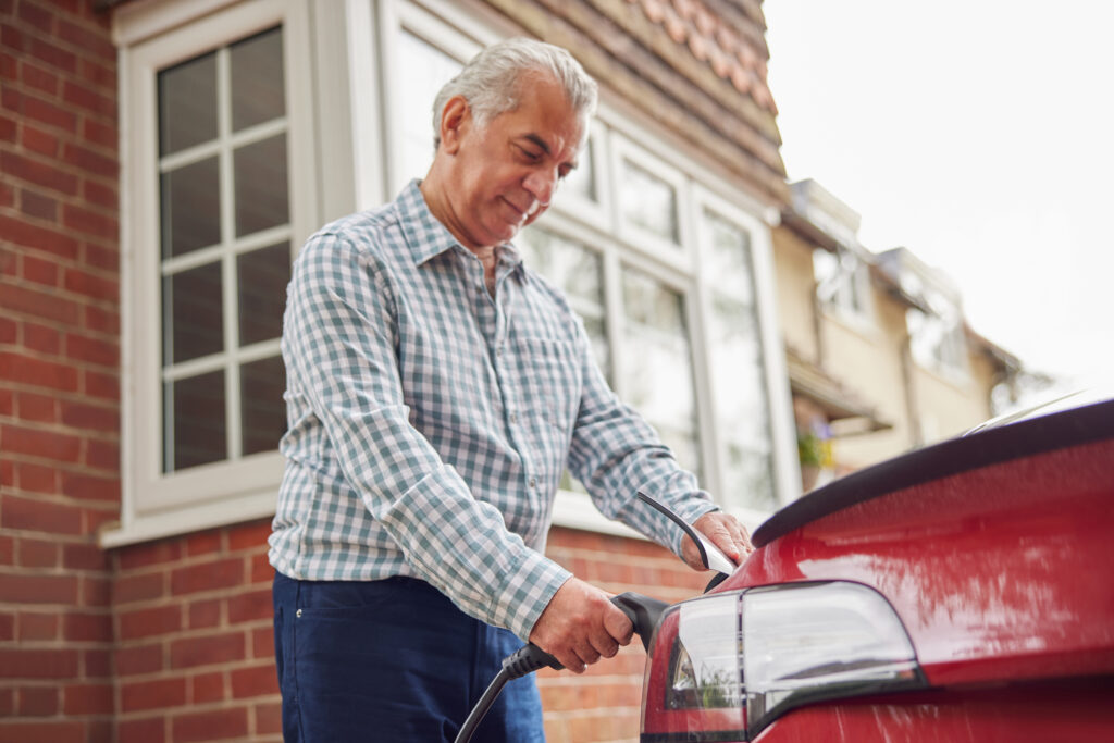 An older man attaches a charging cable to his EV in front of his red brick home for EV-charging-incentives-and-rebates-in-Virginia blog.