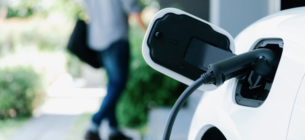 Focus on electric car charging at a home station with blurred man walking in the background for EV-charging-incentives-in-Delaware blog.